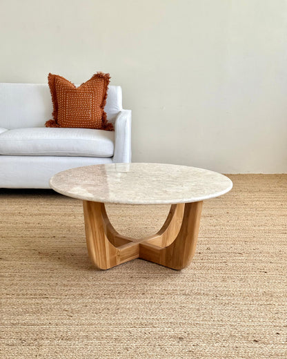 A round coffee table with a light beige stone top and a natural wood base, styled in a living room setting on a woven jute rug with a white sofa in the background.