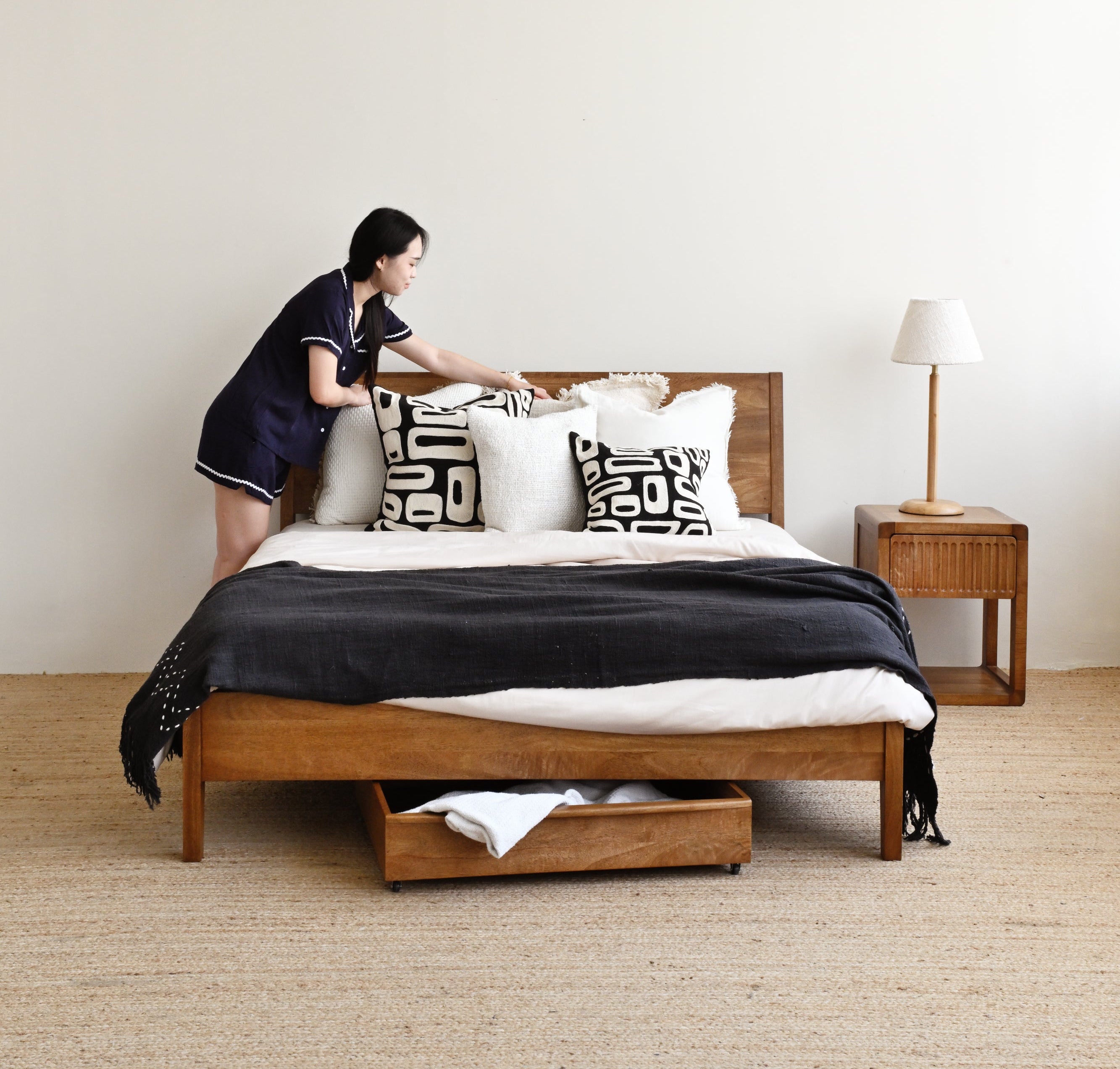 Person making a bed in a modern bedroom with wooden furniture and neutral decor.