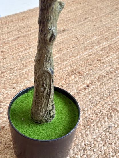 A small artificial tree trunk with textured bark, planted in bright green artificial turf inside a black plastic pot, set on a beige woven rug.