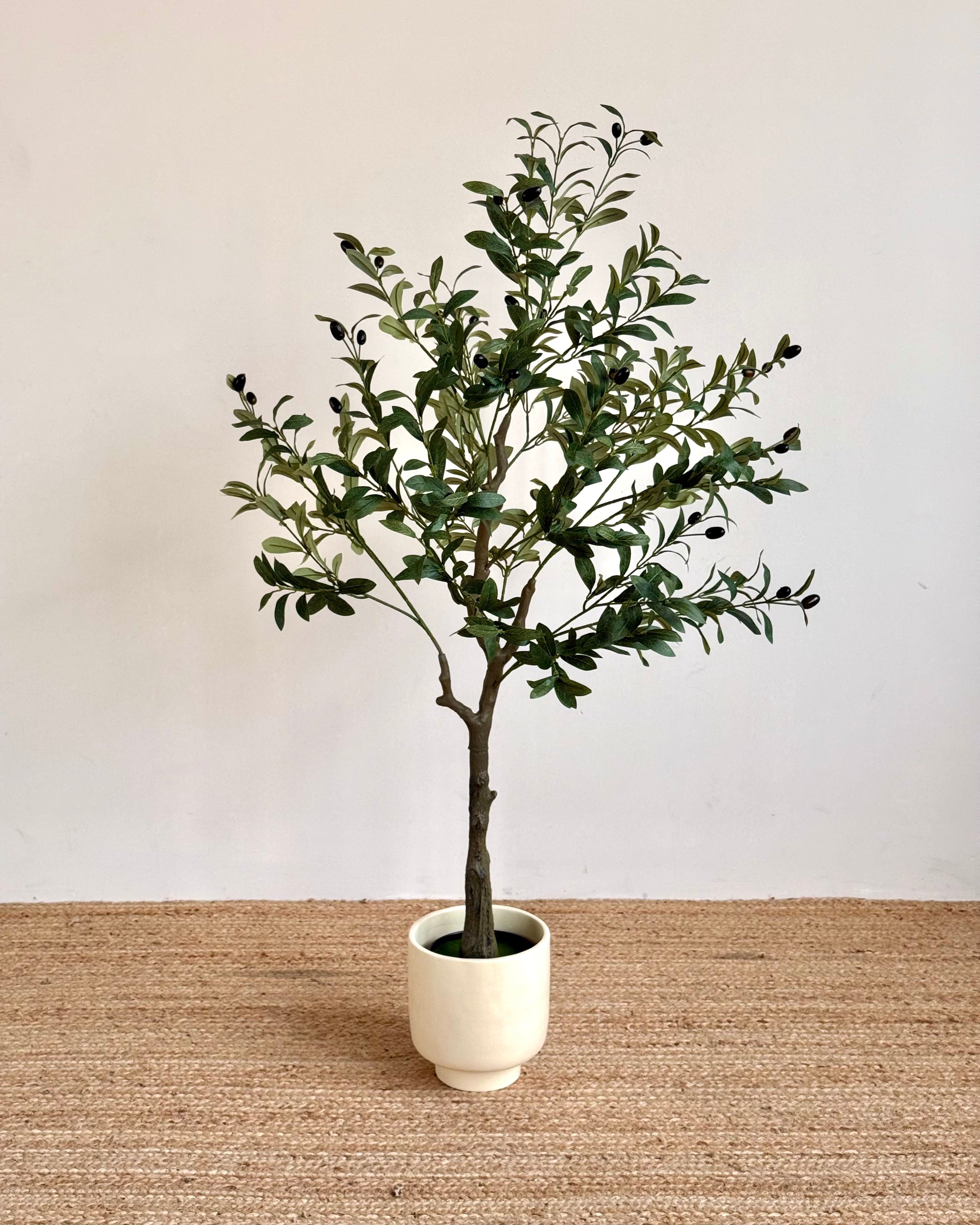 An artificial olive tree with green leaves and small dark berries, potted in a cream-colored ceramic pot and placed on a woven jute rug against a plain off-white wall.