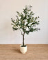An artificial olive tree with green leaves and small dark berries, potted in a cream-colored ceramic pot and placed on a woven jute rug against a plain off-white wall.
