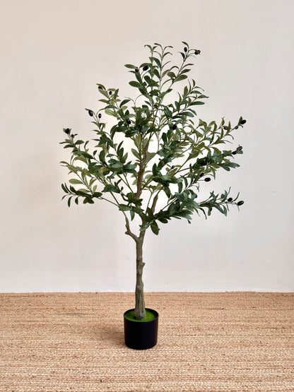 An artificial olive tree with green leaves and small dark olives, planted in a black pot and set on a woven jute rug against a plain off-white wall.