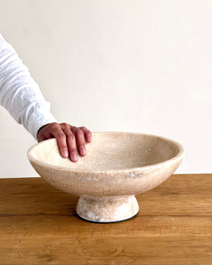 A large, round pedestal bowl made of textured stone or ceramic in a warm beige color, sitting on a wooden table with a person&