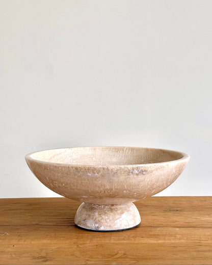 A round pedestal bowl made of textured stone or ceramic in a warm beige and cream color, sitting on a wooden surface against a plain light gray wall.