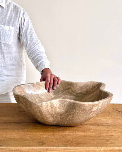 A large organic-shaped wooden bowl with wavy edges sits on a wooden table, with a person&