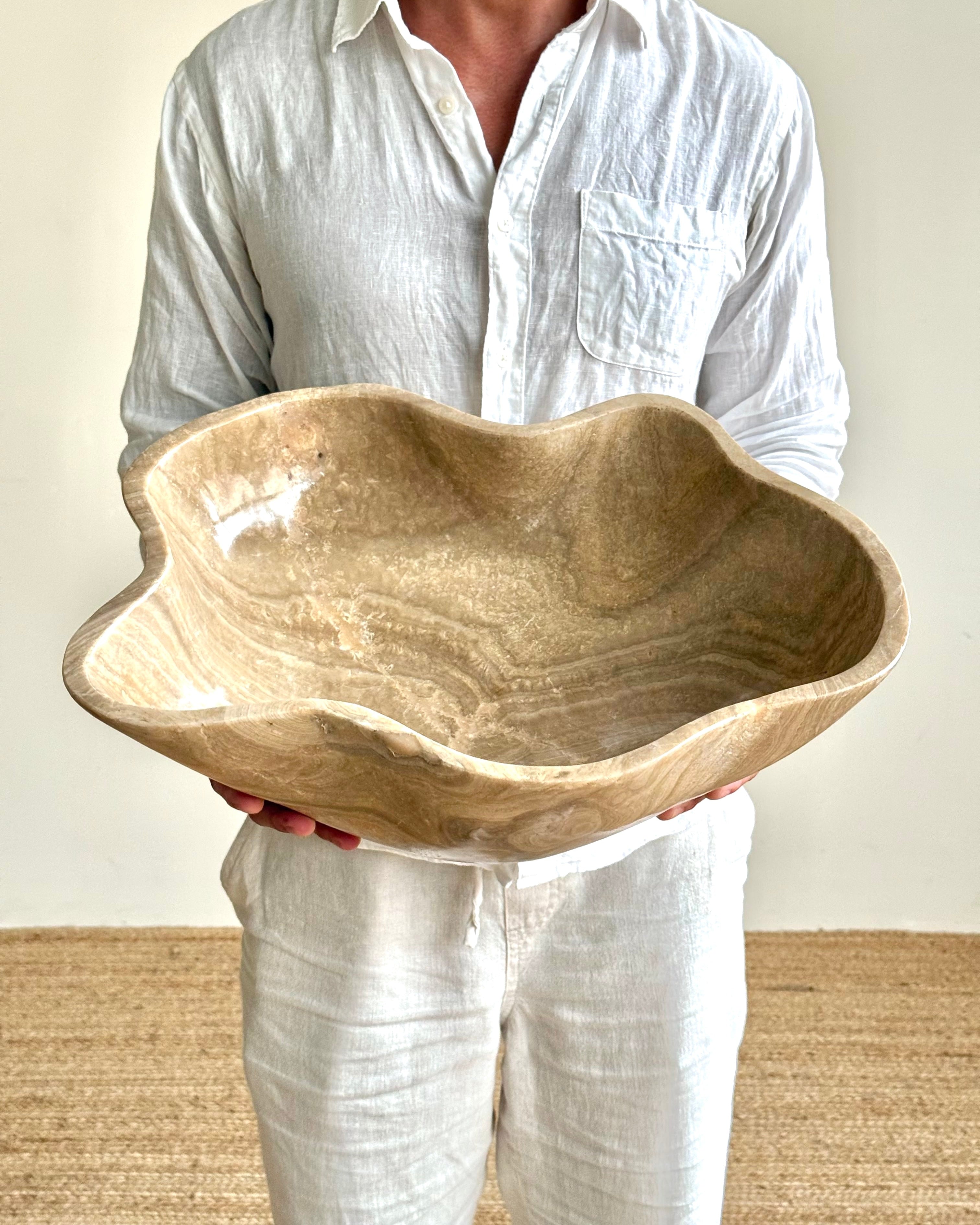 A person wearing a white linen shirt and white pants holds a large, organic-shaped stone bathroom sink basin with a textured beige and brown finish.
