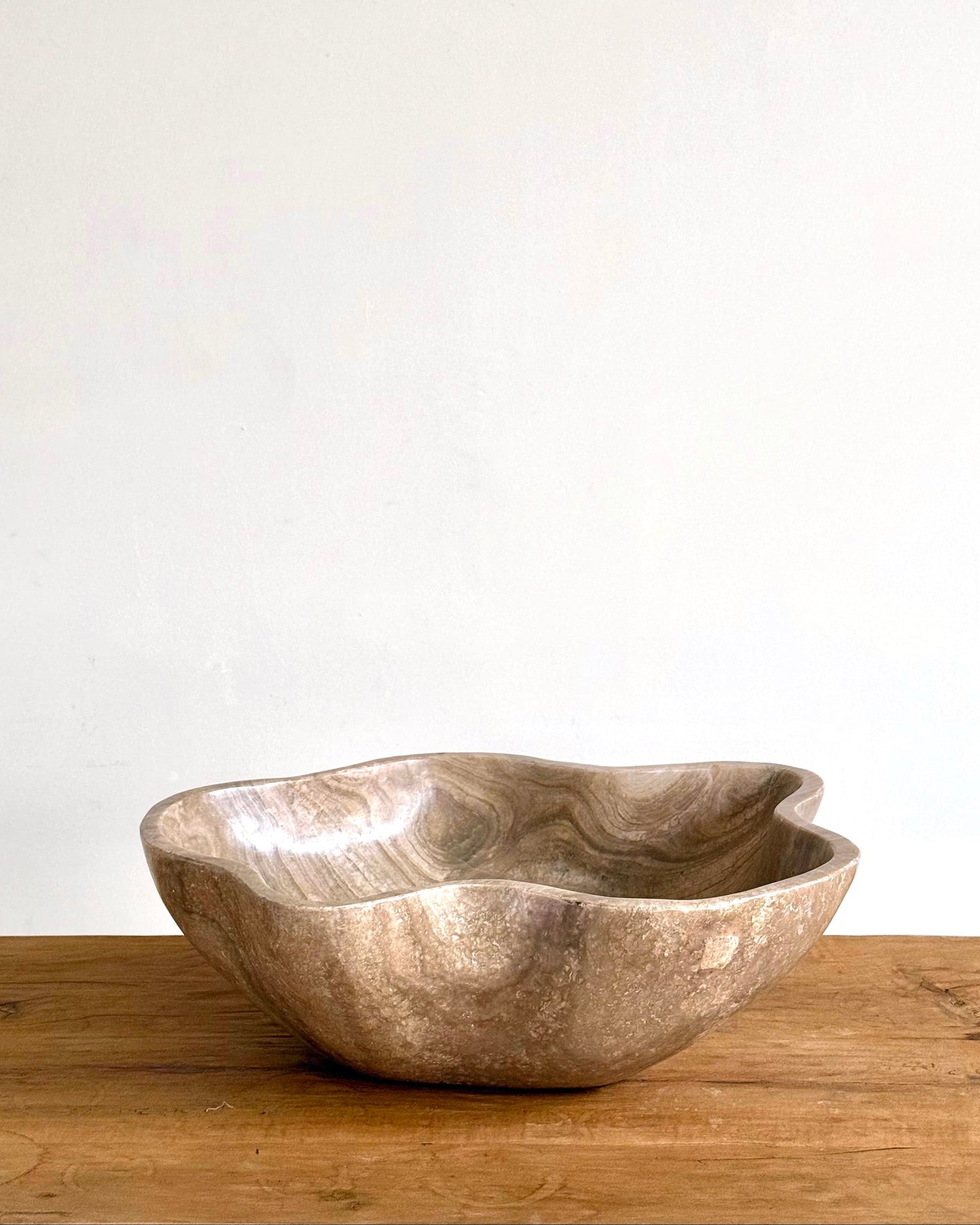 A rustic, organic-shaped stone bowl with a metallic silver and bronze finish, sitting on a wooden surface against a plain off-white wall.