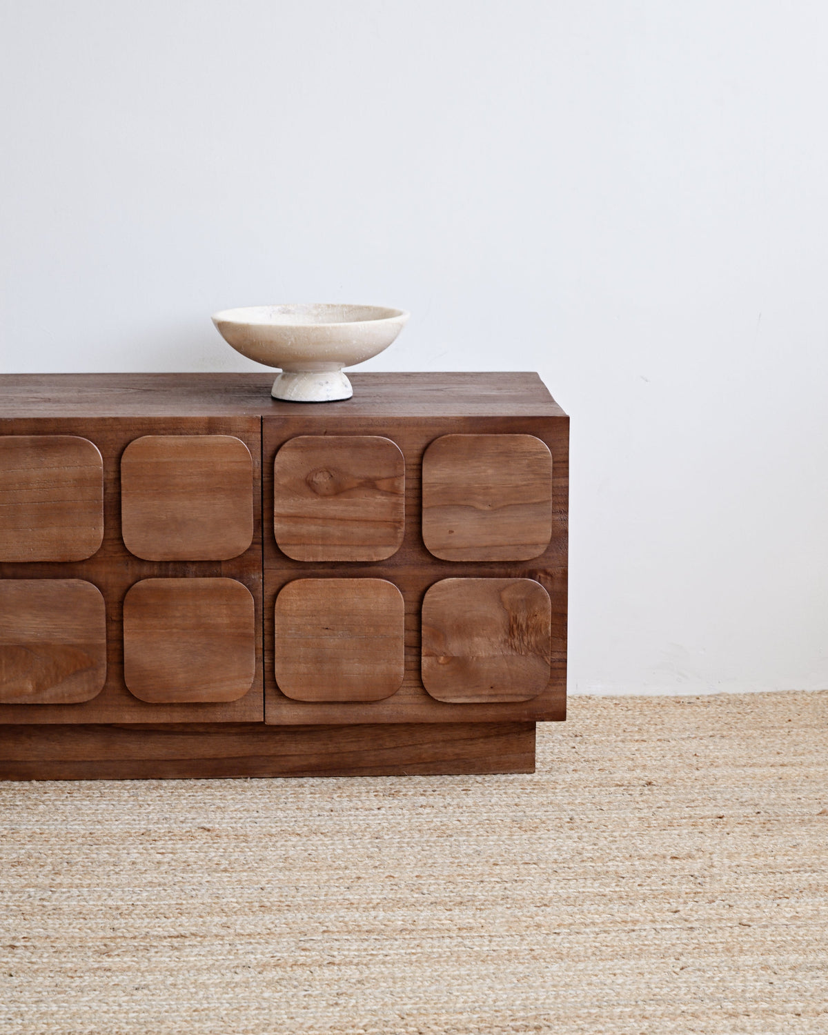 A mid-century modern wooden sideboard with nine rounded square cabinet doors, styled on a natural jute rug with a small white ceramic bowl on top, set against a plain white wall.