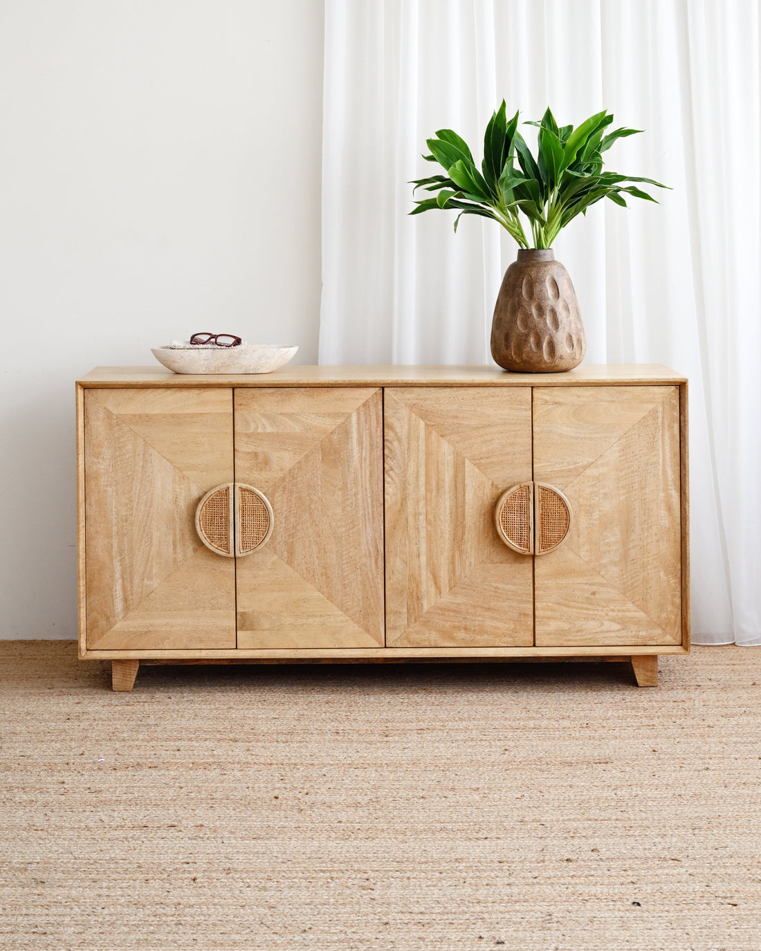 A light natural wood sideboard cabinet with geometric panel detailing and circular carved handles, styled in a bright room with a potted plant and bowl on top.