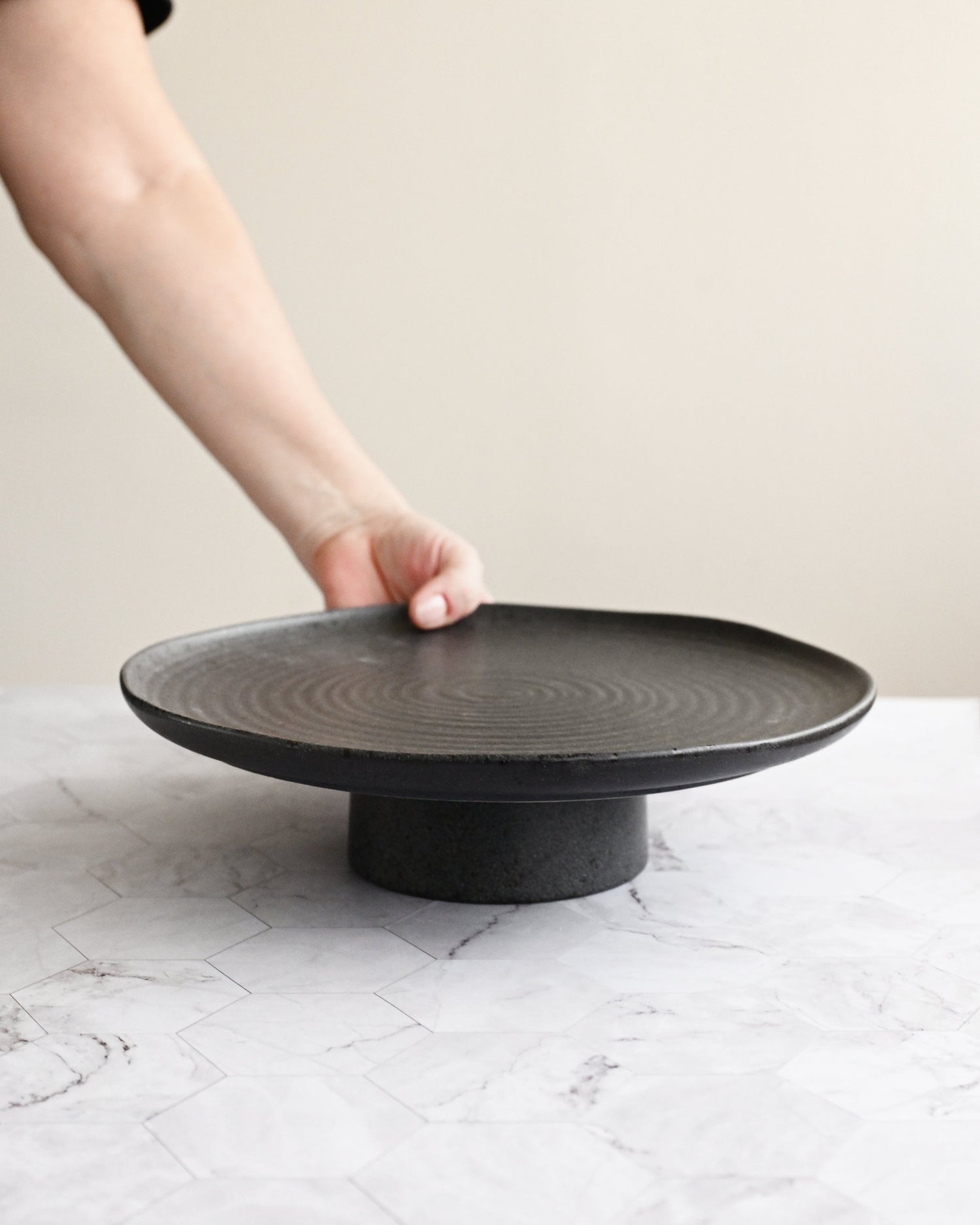 Person holding a black ceramic plate on a matching stand against a neutral background