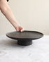 Person holding a black ceramic plate on a matching stand against a neutral background