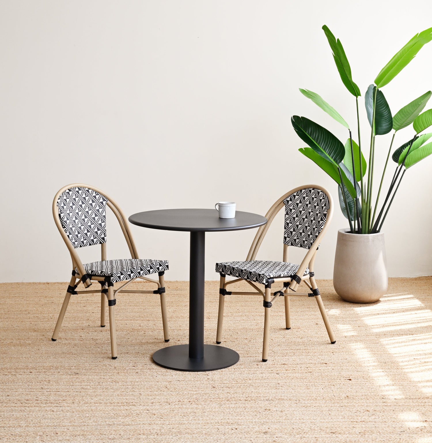 Two chairs with patterned seats and backrests around a round black table, with a potted plant in the background.