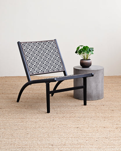 Black chair with patterned backrest next to a small concrete table with a plant on a beige carpet.