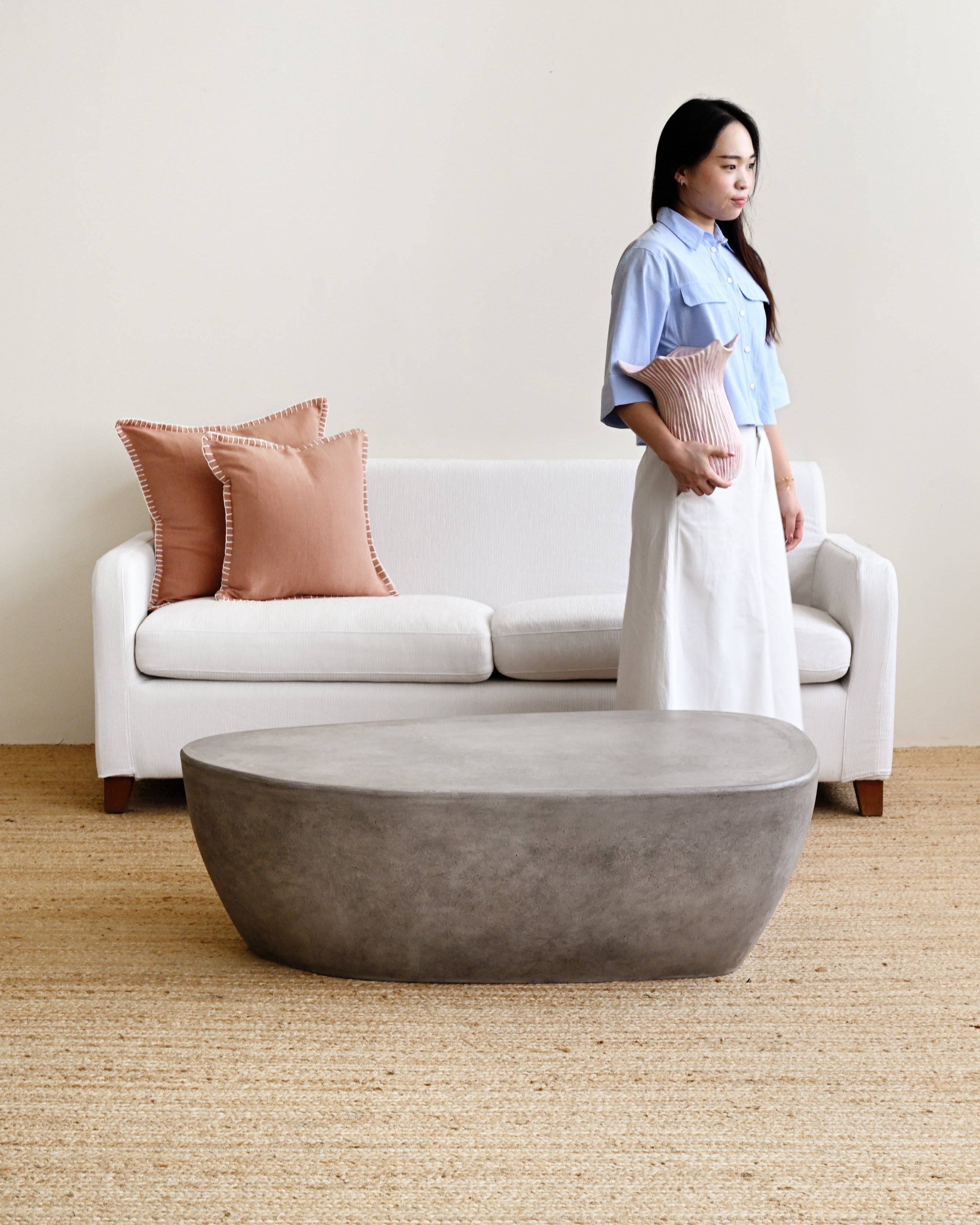 Woman standing in a living room with a white sofa, pink pillows, and a concrete coffee table.