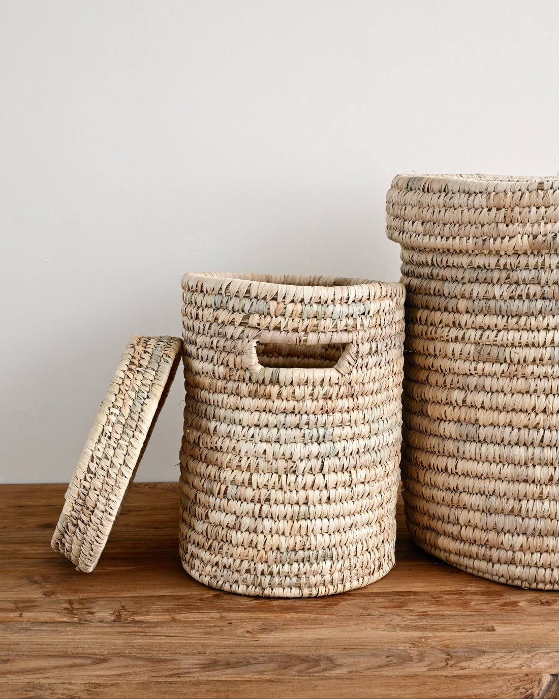 Three woven seagrass storage baskets in natural beige and cream tones, including two cylindrical baskets with cutout handles and a rectangular basket with a lid, displayed on a rustic wooden surface against a plain off-white wall.