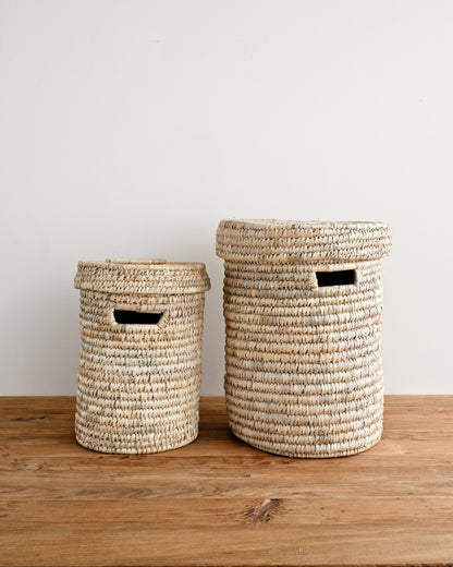 Two woven natural-fiber storage baskets with black handles, one small and one large, displayed on a wooden surface against a plain light gray wall.