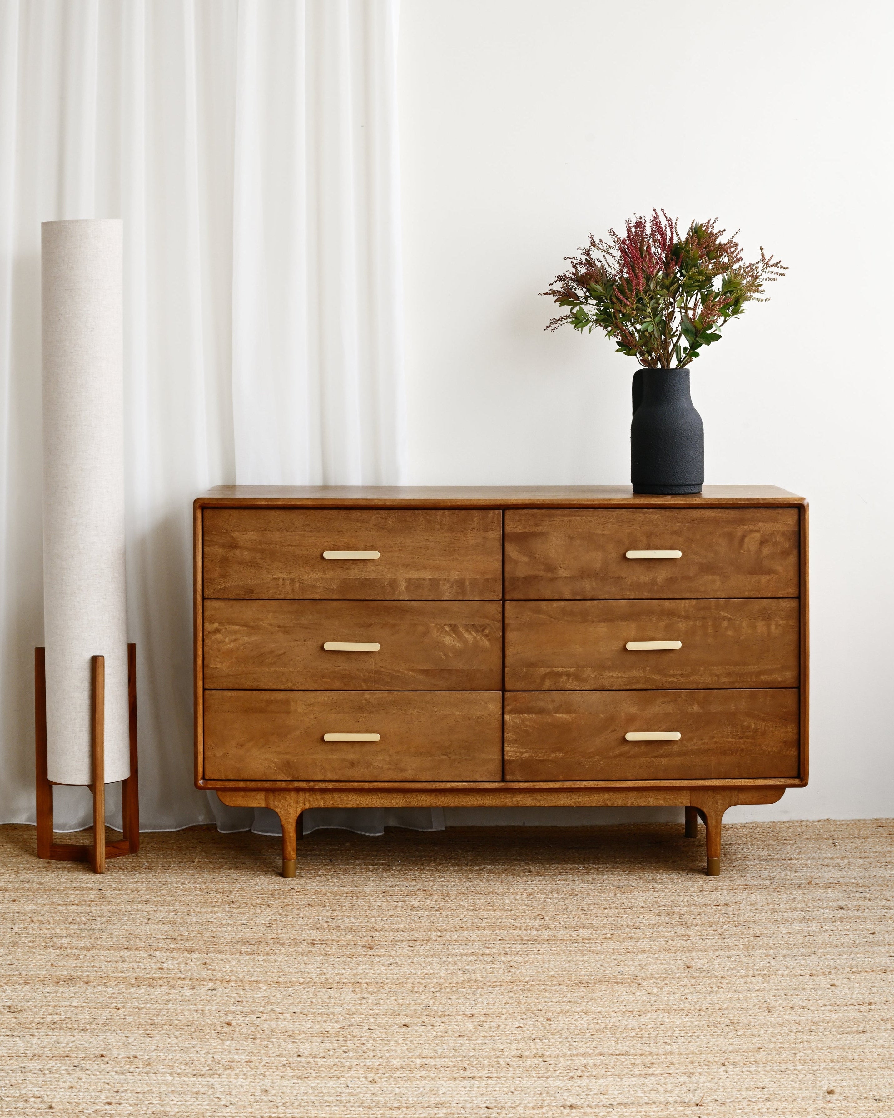 Wooden dresser with a vase of flowers and a lamp in a minimalistic room.