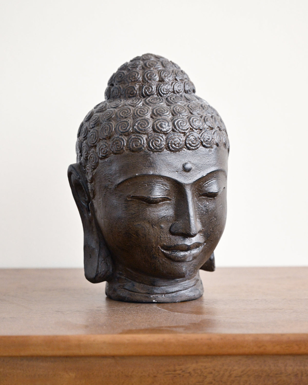 A dark bronze Buddha head sculpture displayed on a wooden surface against a plain off-white background.