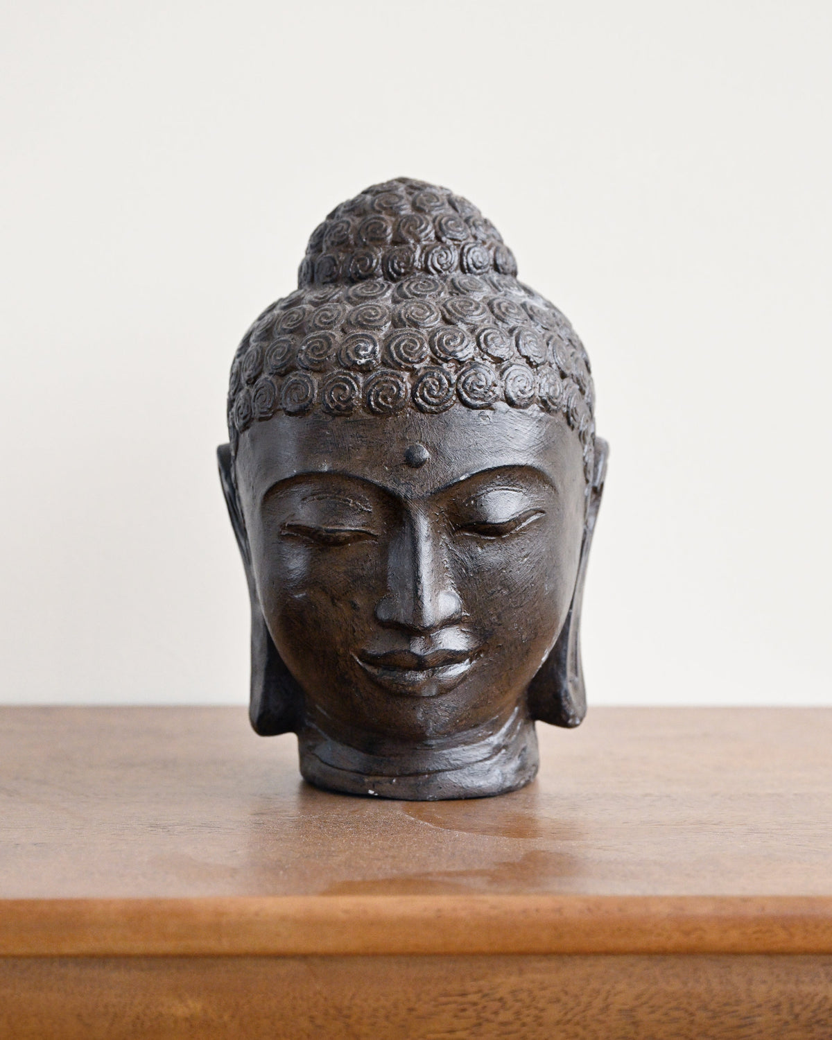 A dark bronze Buddha head sculpture displayed on a wooden surface against a plain off-white background.