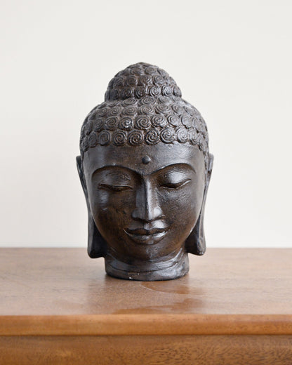 A dark bronze Buddha head sculpture displayed on a wooden surface against a plain off-white background.