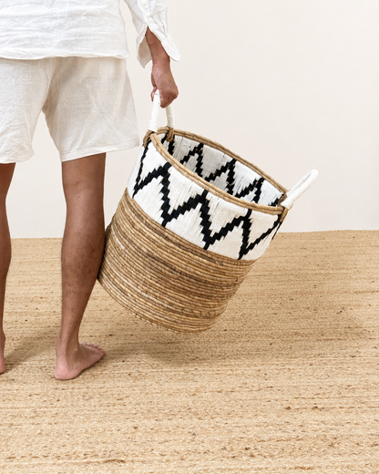 Person holding a woven basket with a black and white pattern on a wooden floor.