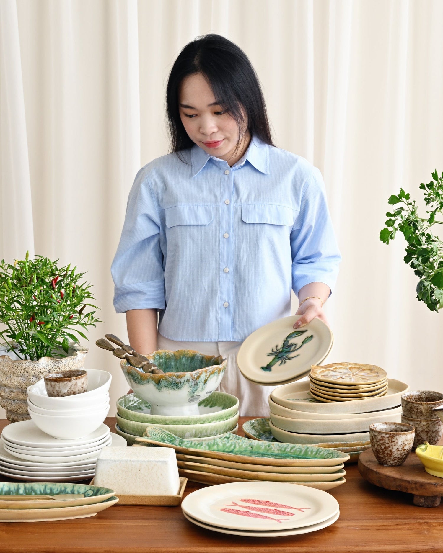 Woman arranging ceramic plates and bowls on a wooden table with plants in the background.