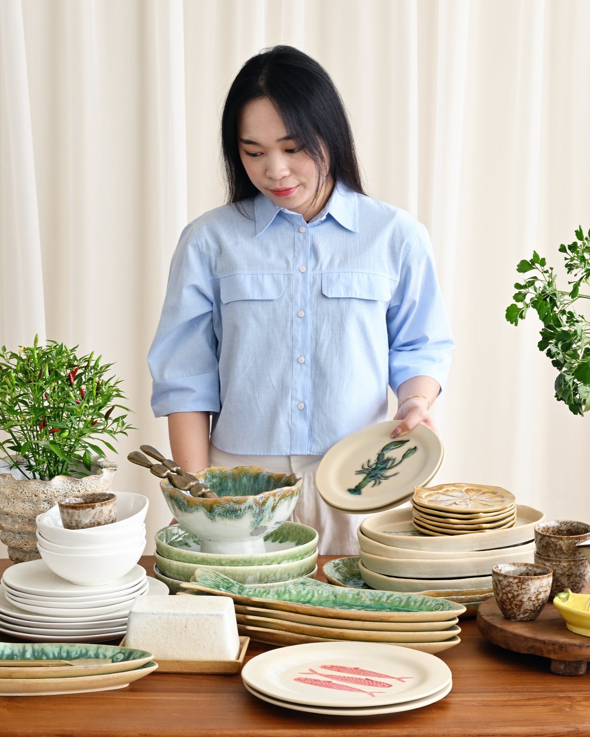 Woman arranging ceramic plates and bowls on a wooden table with plants in the background.