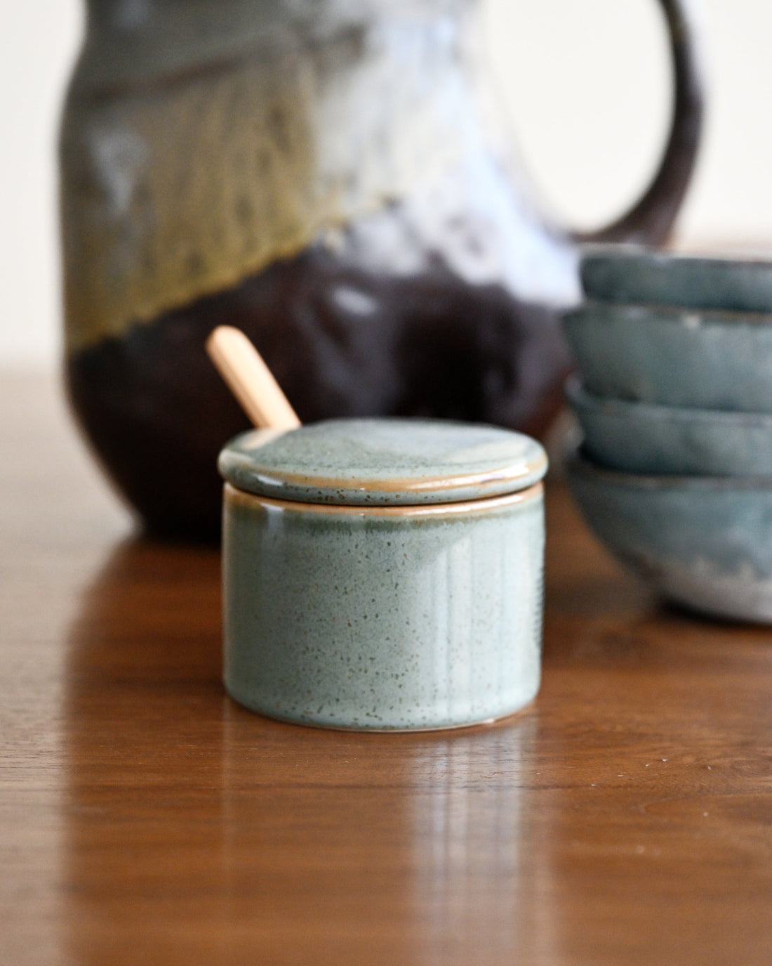 A small speckled ceramic jar with a matching lid and a wooden spoon, displayed on a wooden table with a blurred ceramic pitcher and bowls in the background.