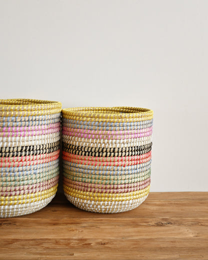 Two woven storage baskets with horizontal multicolor stripes in yellow, pink, orange, black, and white, sitting on a wooden surface against a plain light gray wall.