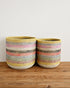 Two handwoven cylindrical baskets with horizontal multicolor stripes in yellow, pink, blue, black, and white, displayed on a wooden surface against a plain white wall.