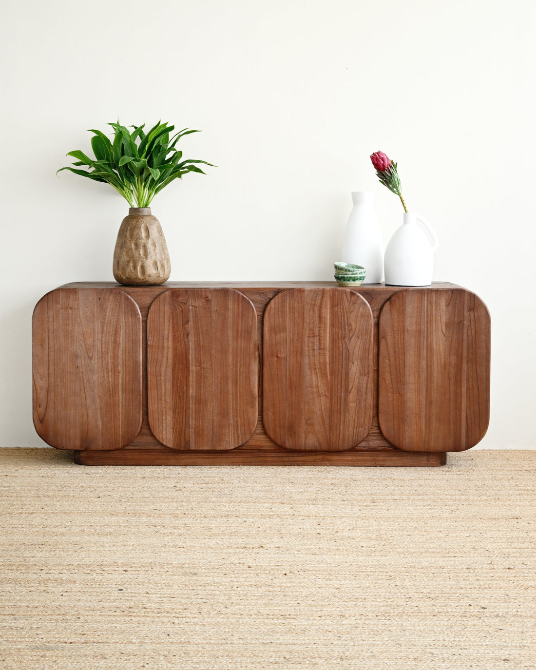 A mid-century modern wooden sideboard cabinet with rounded panel doors, styled with a potted green plant and white ceramic vases on top, set against a plain off-white wall and light beige woven rug.