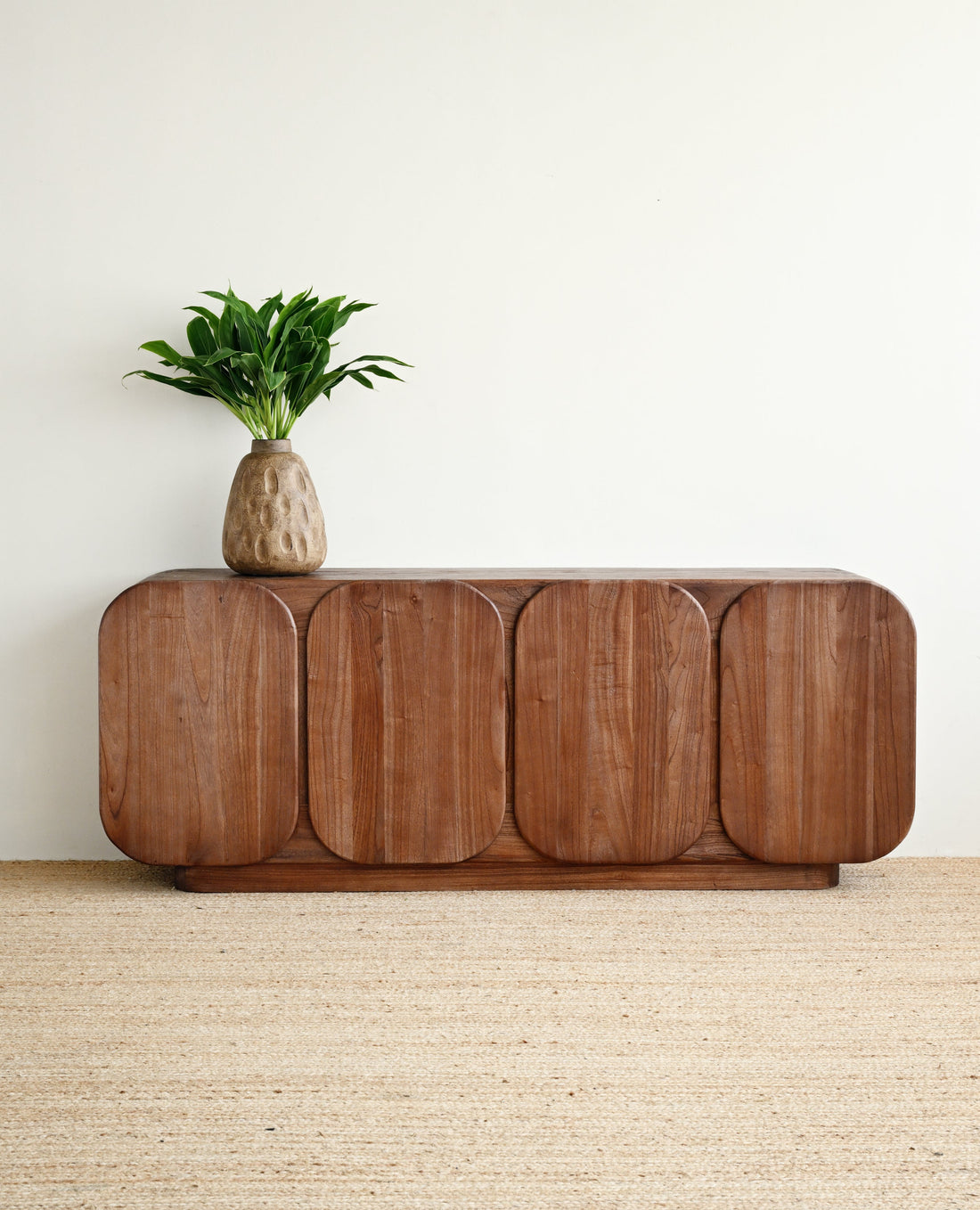 A mid-century modern wooden credenza with rounded edges and four arched door panels, styled with a small potted green plant on top, set against a plain off-white wall and a textured beige jute rug.