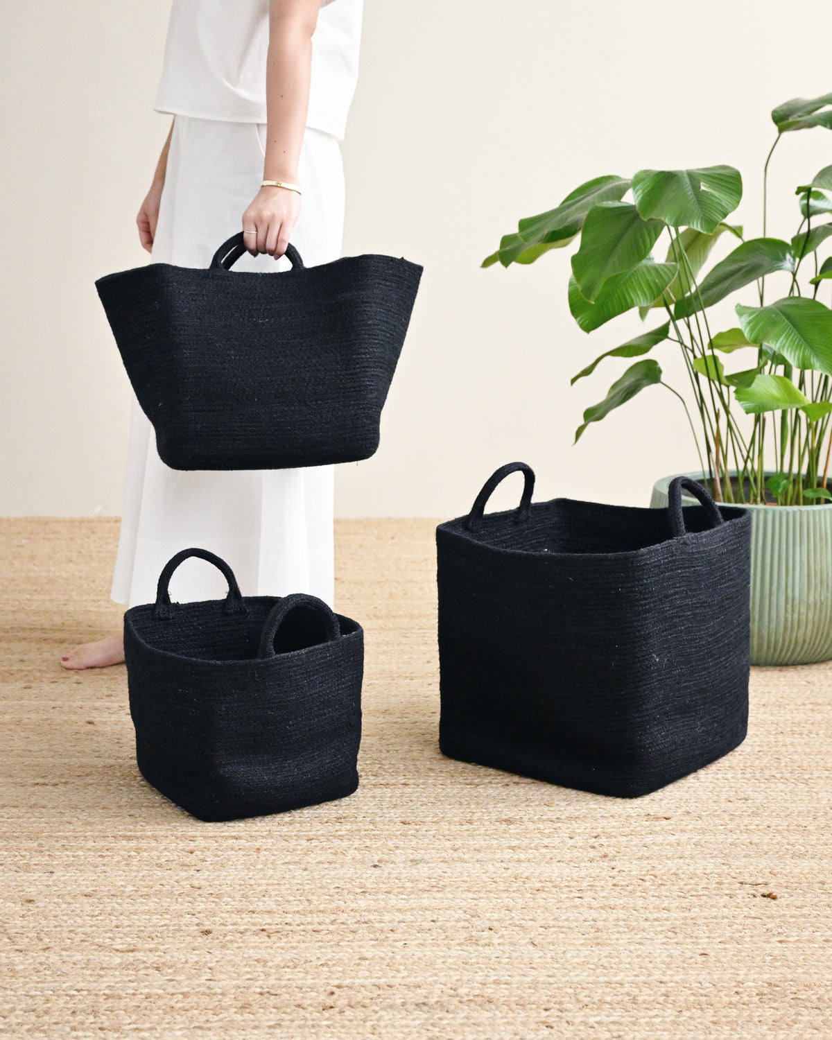 Three black woven storage baskets with handles are displayed on a beige jute rug, with a woman in a white outfit holding one of the baskets and a green potted plant in the background.