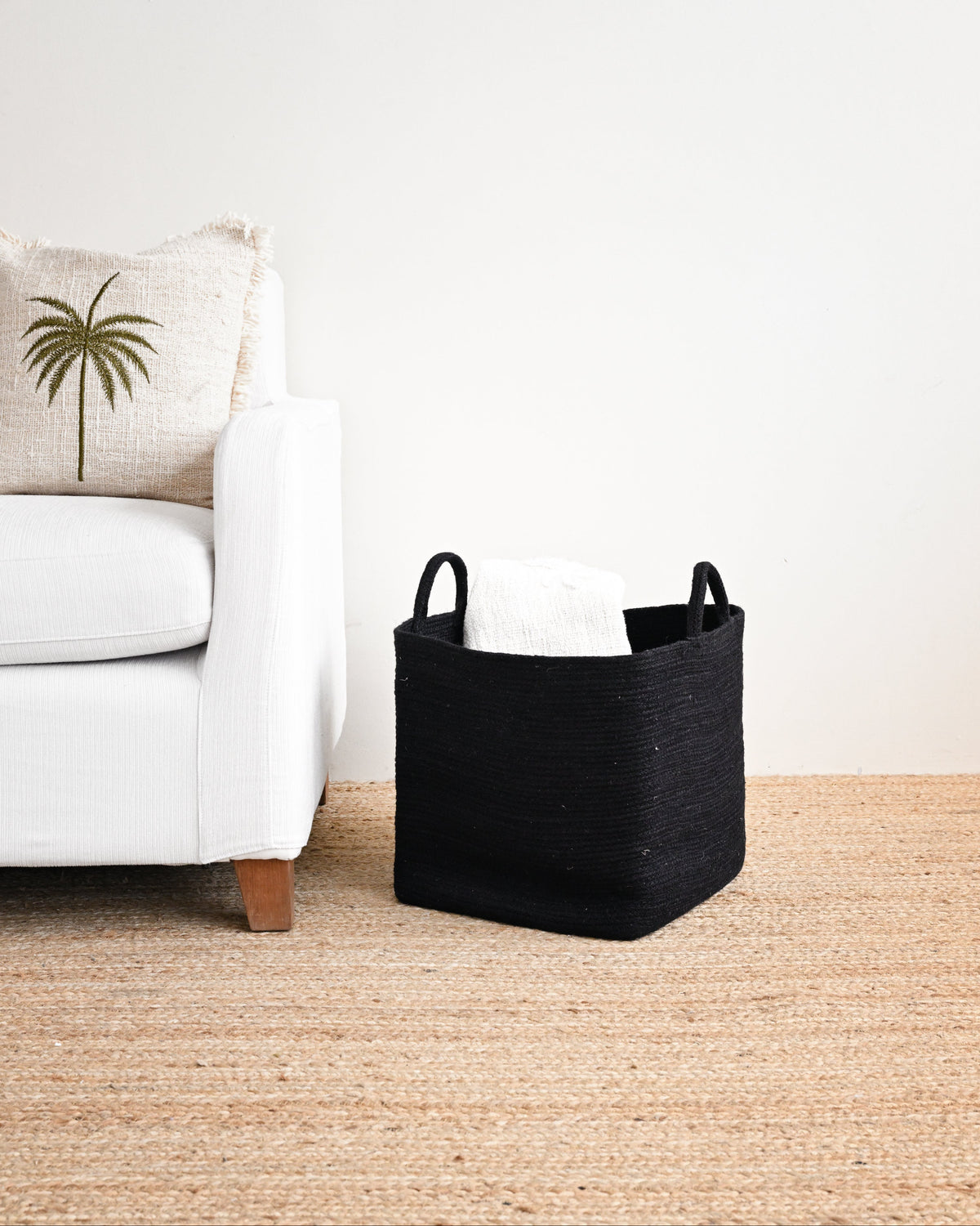A black woven storage basket with two handles sits on a natural jute rug next to a white armchair with a palm tree pillow.