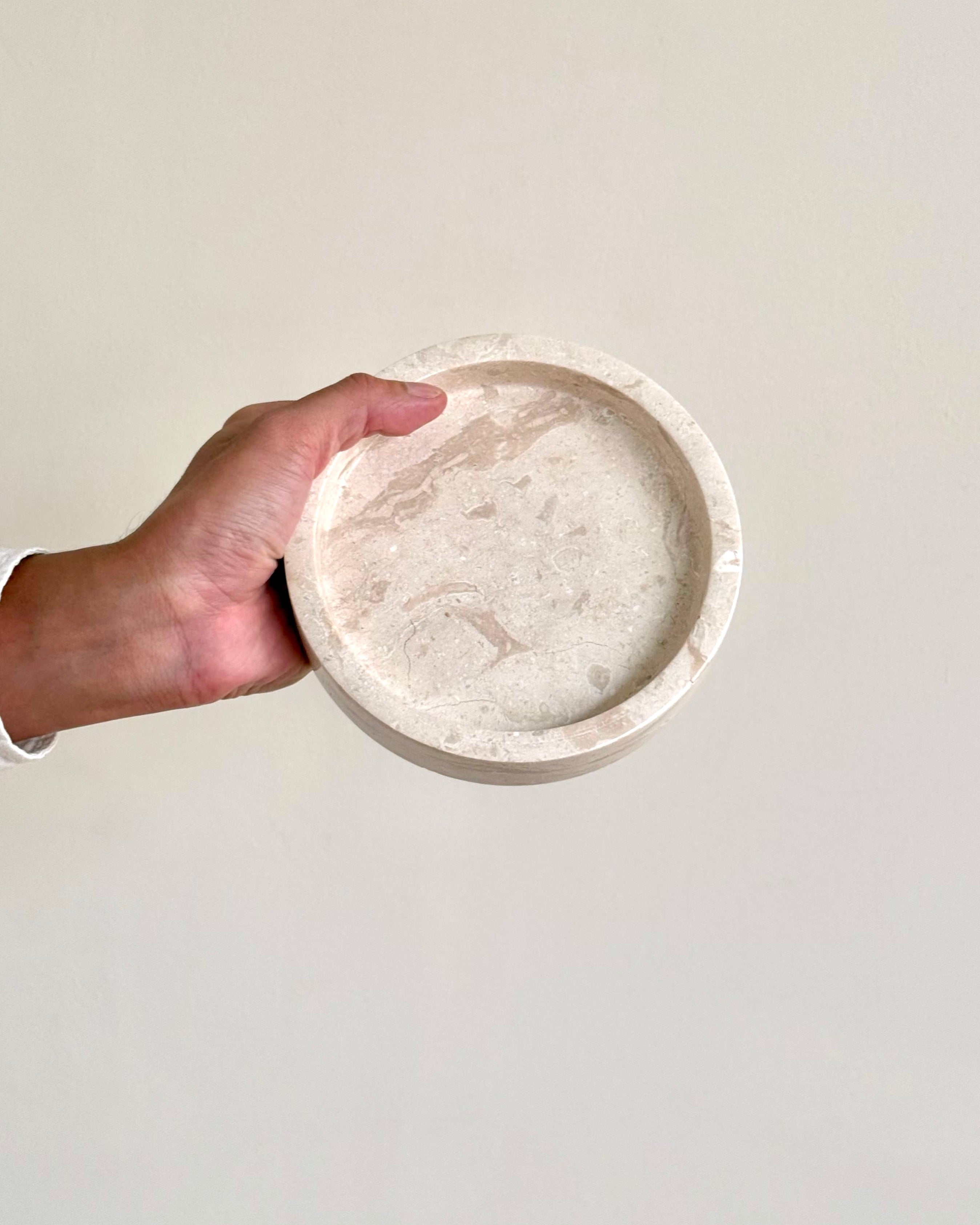 A hand holding a round, shallow stone tray with a light beige and off-white marbled texture against a plain, warm off-white wall.