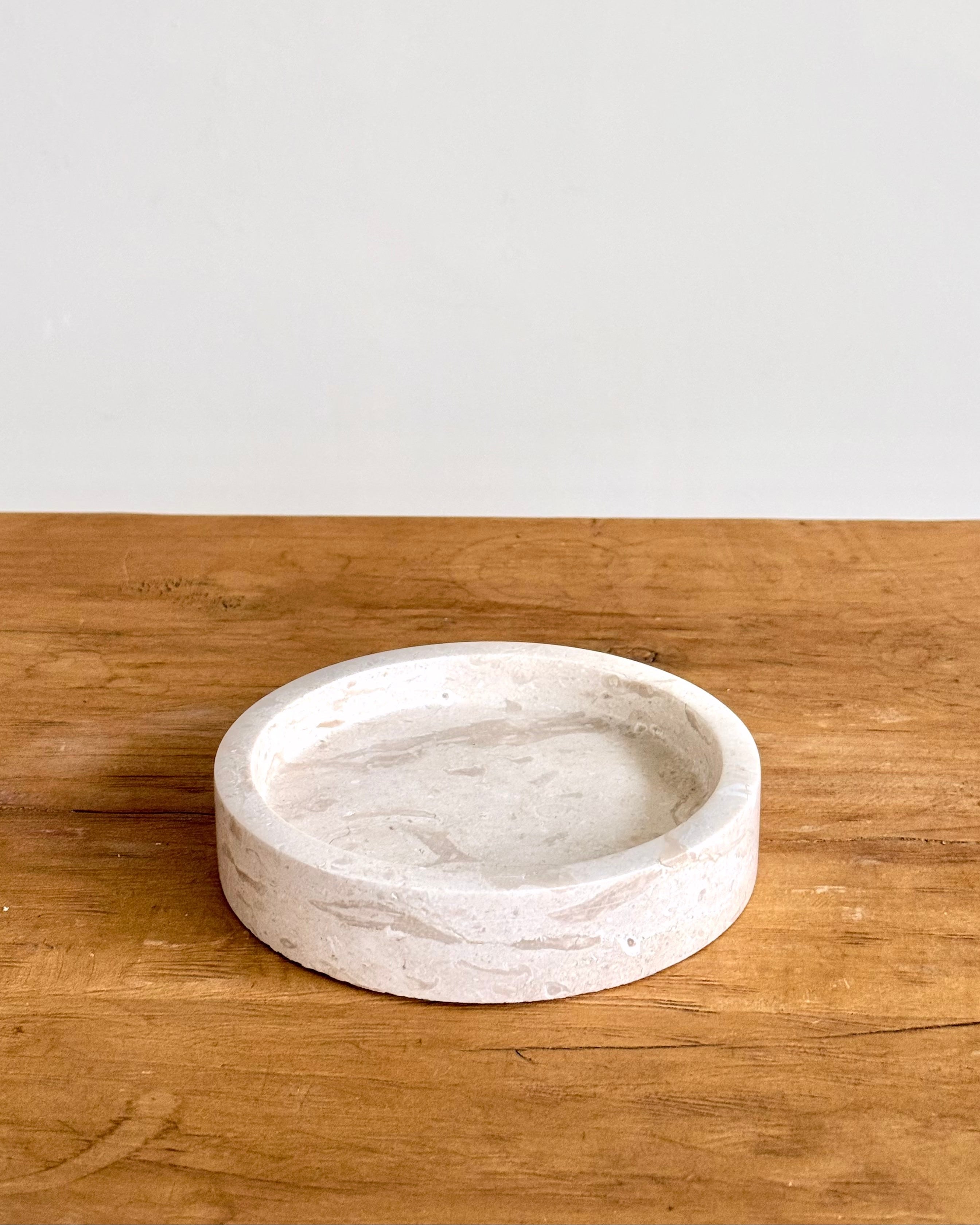 A round, shallow stone tray with a textured, natural finish, displayed on a rustic wooden surface against a plain white wall.