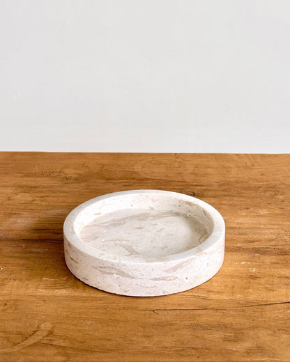 A round, shallow stone tray with a textured, natural finish, displayed on a rustic wooden surface against a plain white wall.
