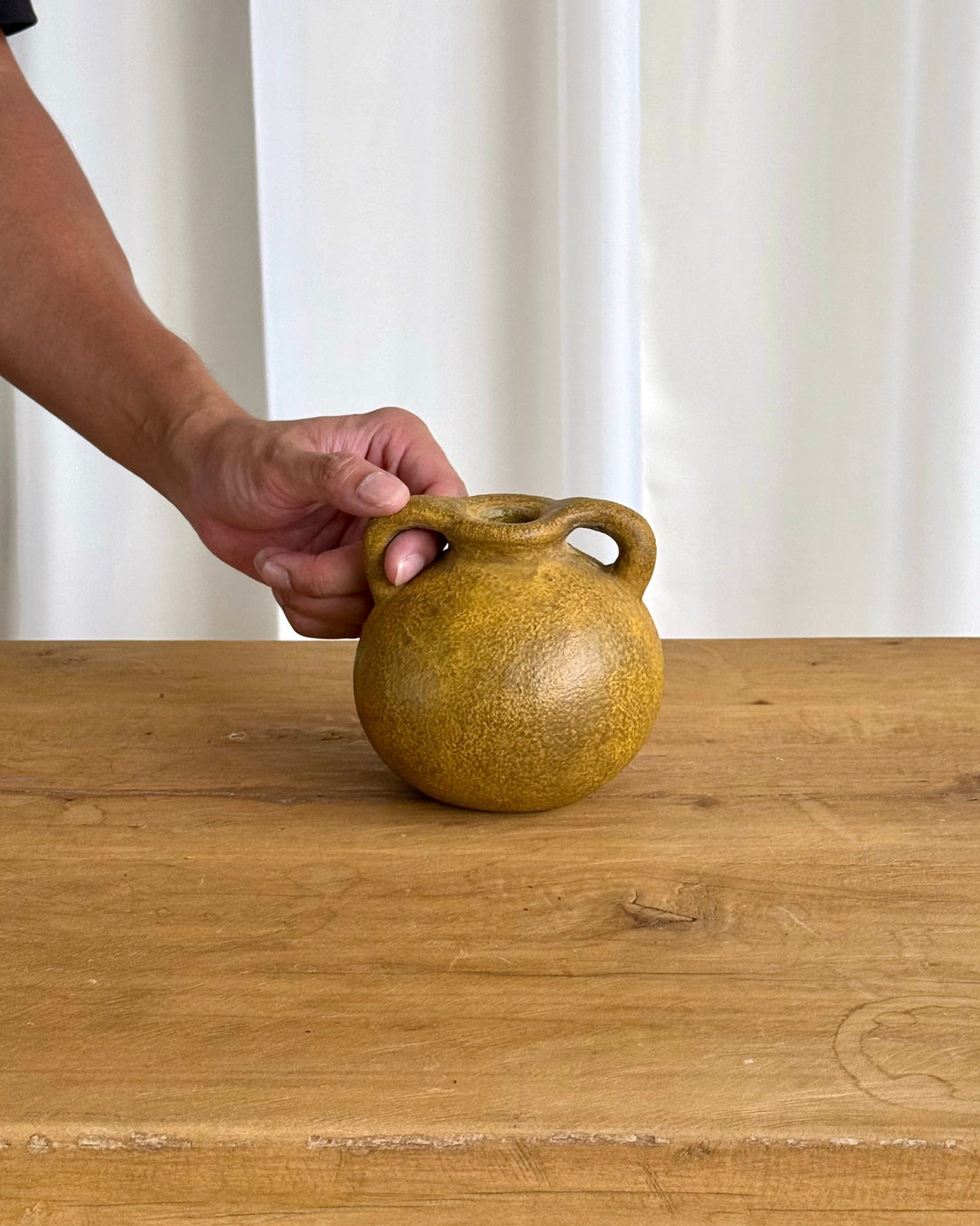 A small round ceramic jug with a single loop handle, shown on a wooden table with a person&