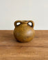 A small round ceramic vase with two loop handles, featuring a textured speckled glaze in earthy brown and ochre tones, set on a wooden tabletop against a plain light gray wall.