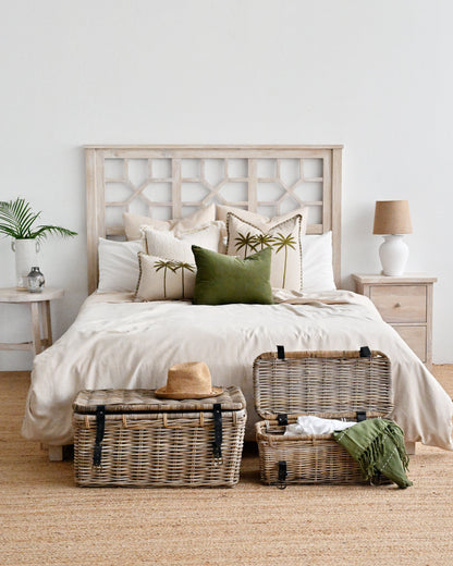 A styled bedroom scene with a bed dressed in cream bedding and decorative pillows, flanked by two small side tables with lamps, and wicker storage trunks on the floor in front of the bed.