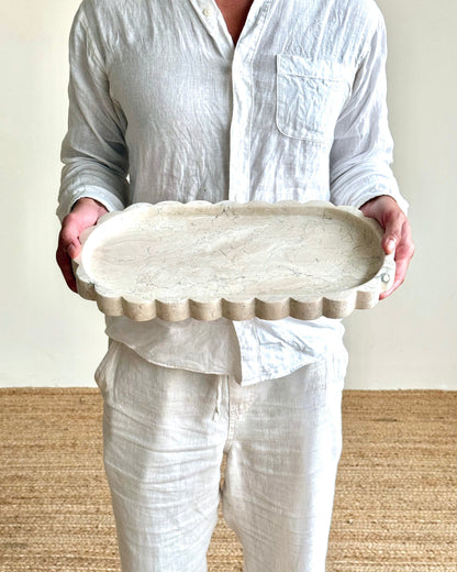 A person wearing a white linen shirt and pants holds a scalloped oval ceramic serving tray against a neutral indoor background.