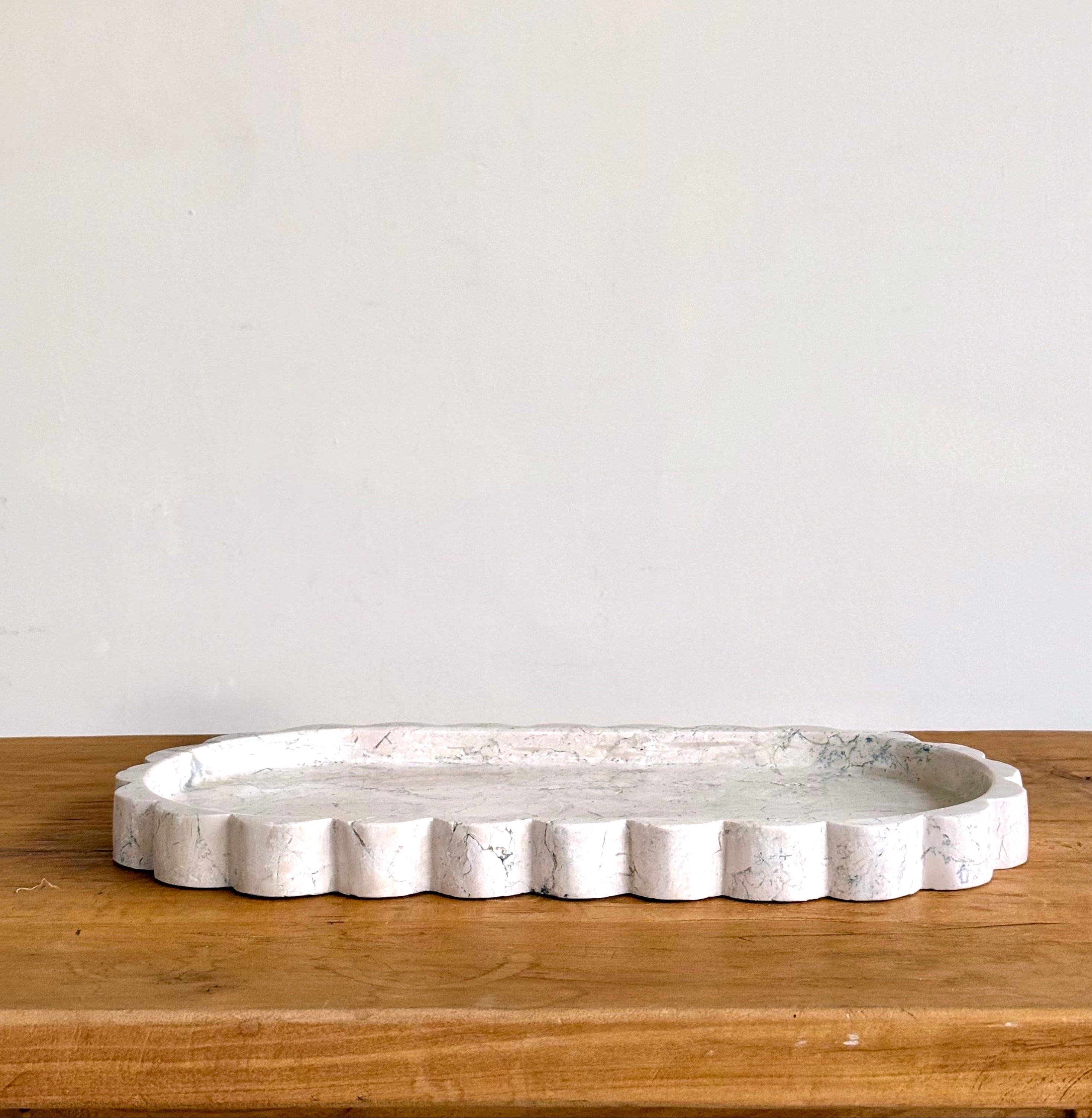 An oval white tray with a scalloped edge and a distressed, stone-like texture, resting on a wooden surface against a plain off-white wall.