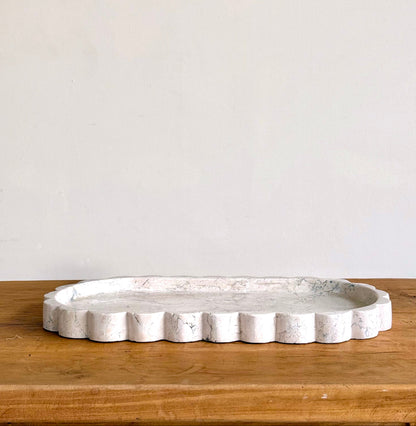 An oval white tray with a scalloped edge and a distressed, stone-like texture, resting on a wooden surface against a plain off-white wall.