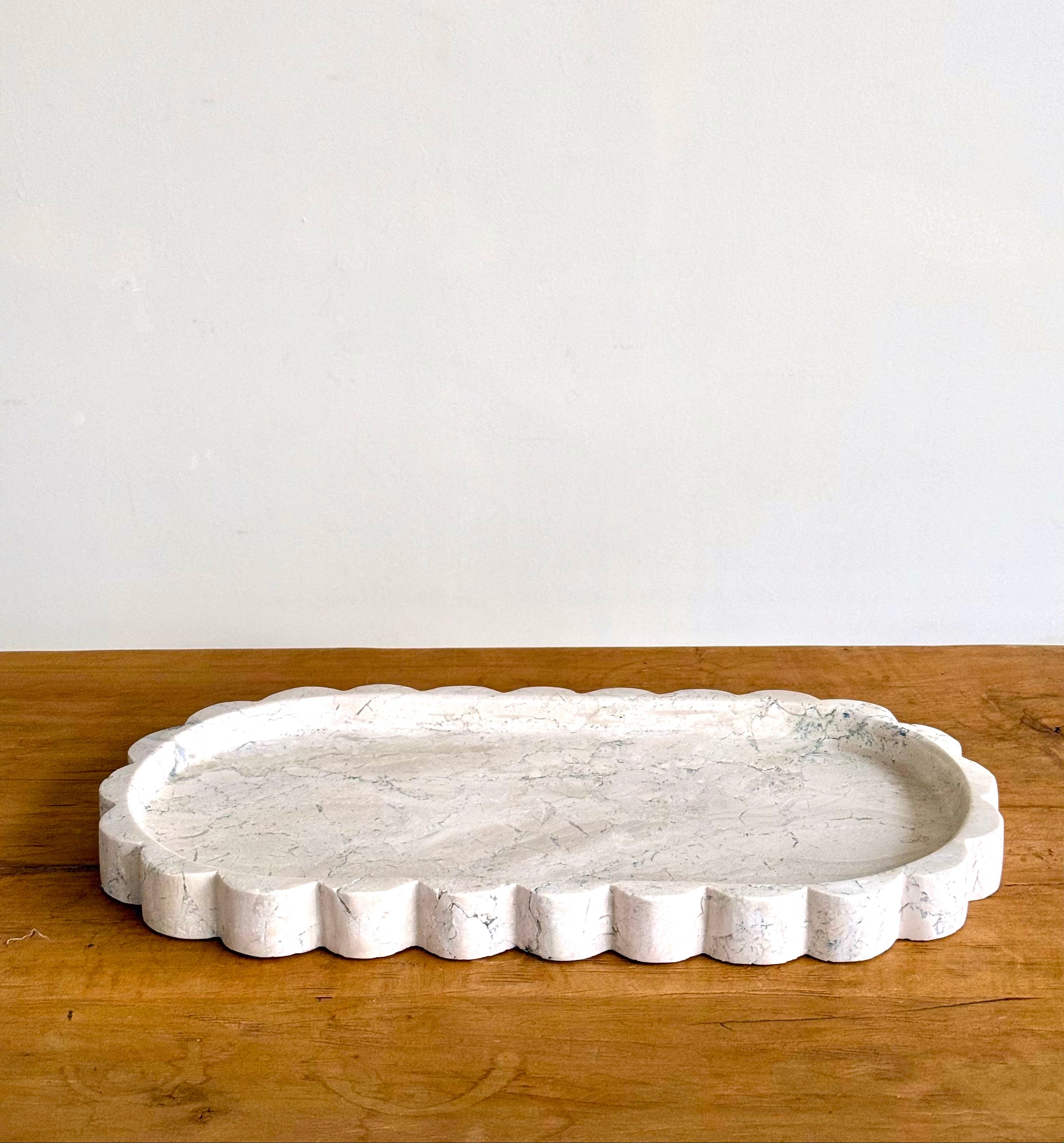 A scalloped white stone serving tray with a textured, stone-like surface, resting on a wooden tabletop against a plain off-white wall.