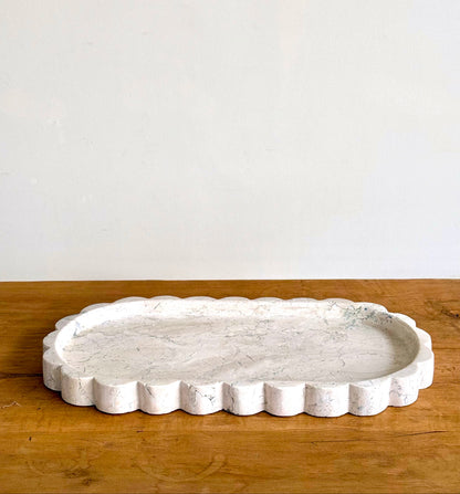 A scalloped white stone serving tray with a textured, stone-like surface, resting on a wooden tabletop against a plain off-white wall.