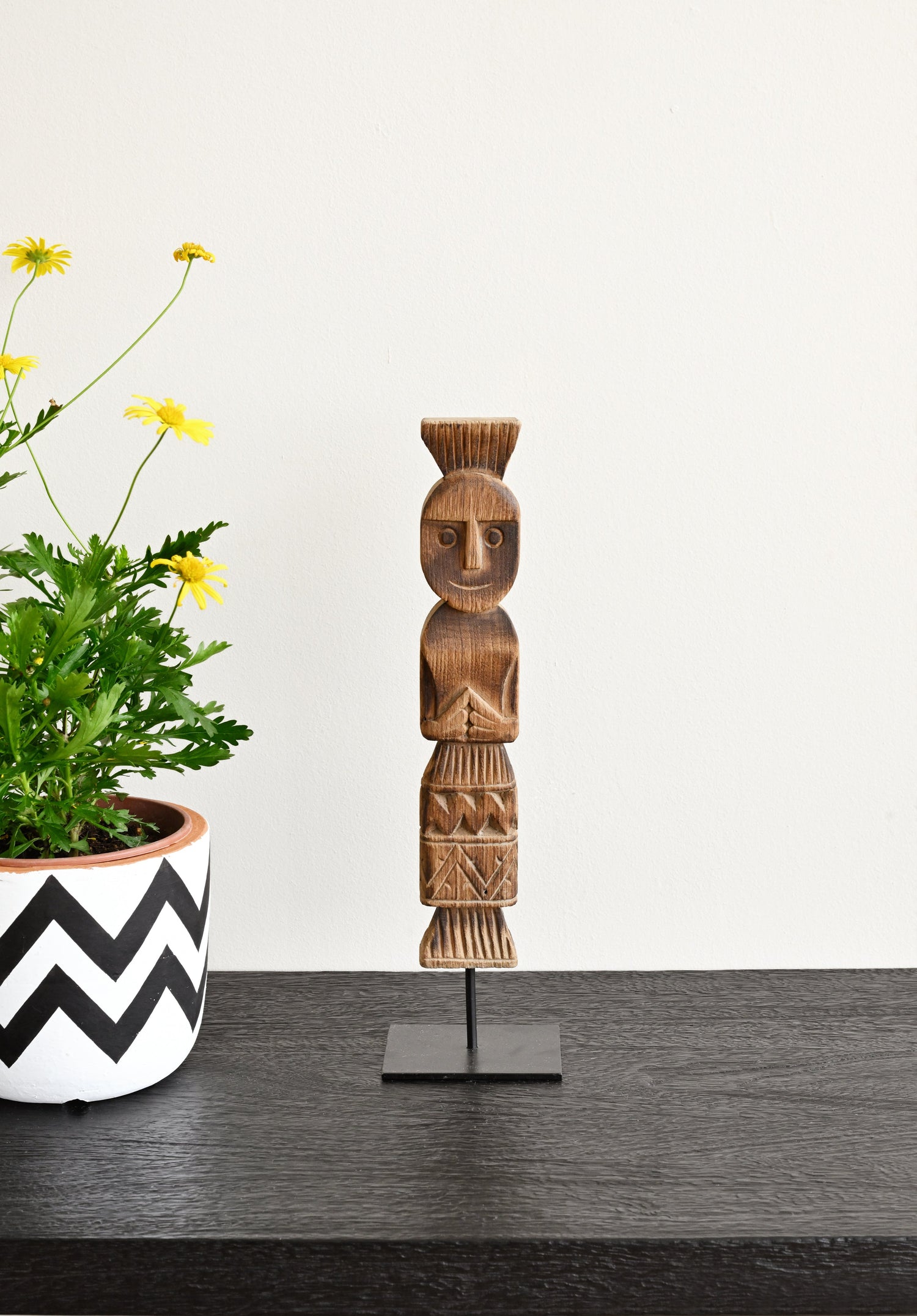 Wooden figurine on a stand next to a potted plant with a white wall background