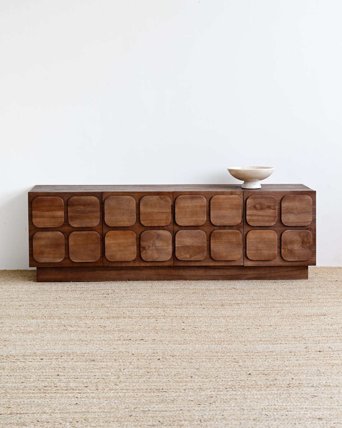 A mid-century modern wooden sideboard with a grid of rounded square doors, shown on a light beige jute rug against a white wall with a small ceramic bowl on top.