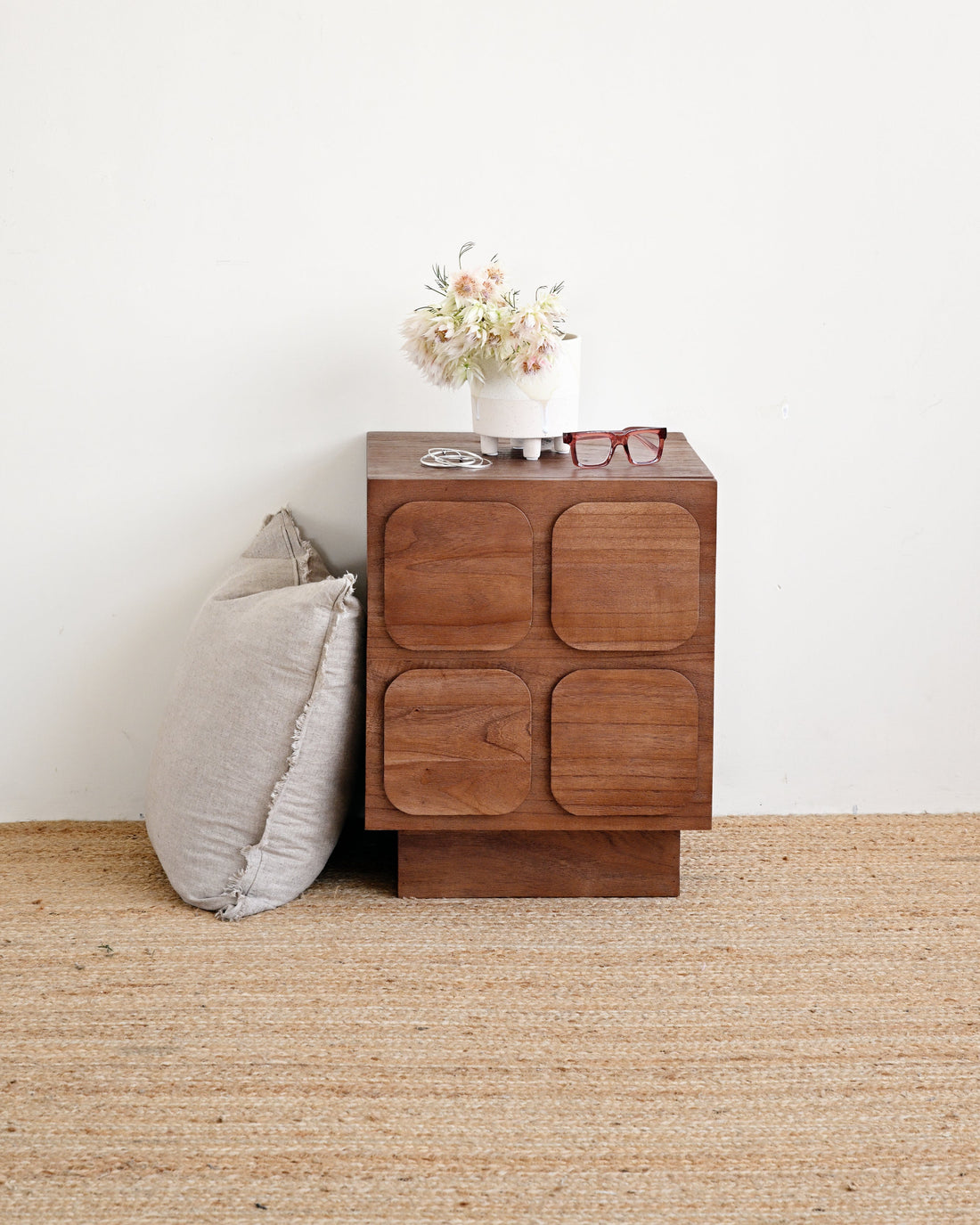 A mid-century modern wooden nightstand with four square recessed drawer fronts, styled next to a gray pillow on a woven jute rug against a white wall.