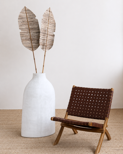 White vase with dried leaves next to a brown woven chair on a beige carpet.