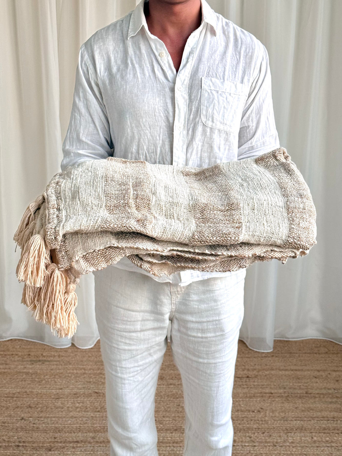 A person wearing a white linen shirt and white pants holds a folded beige and cream woven throw blanket with tassels in front of a white curtain background.